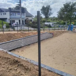 Wykopany teren z częściowo wzniesioną betonową konstrukcją oporową z bloczków, widoczne metalowe słupki i przygotowany teren pod budowę, w tle dom jednorodzinny i błękitne niebo.