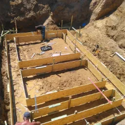 Przygotowanie wykopu pod fundamenty z deskowaniem z desek i ziemnym nasypem w tle, dwóch pracowników w trakcie prac budowlanych, narzędzia widoczne w środku wykopu.