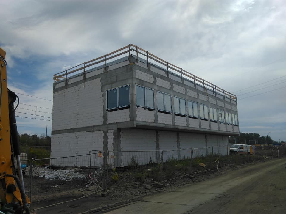 Dwukondygnacyjny budynek w stanie surowym zamkniętym, wzniesiony z białych bloczków, z widocznymi oknami i drewnianą barierką na dachu, na tle pochmurnego nieba i z żółtą koparką widoczną z lewej...