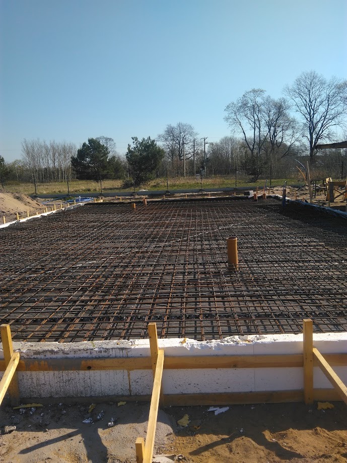 Przygotowanie zbrojenia pod płytę fundamentową domu jednorodzinnego z widoczną izolacją termiczną i rurami instalacyjnymi, na tle drzew i błękitnego nieba.