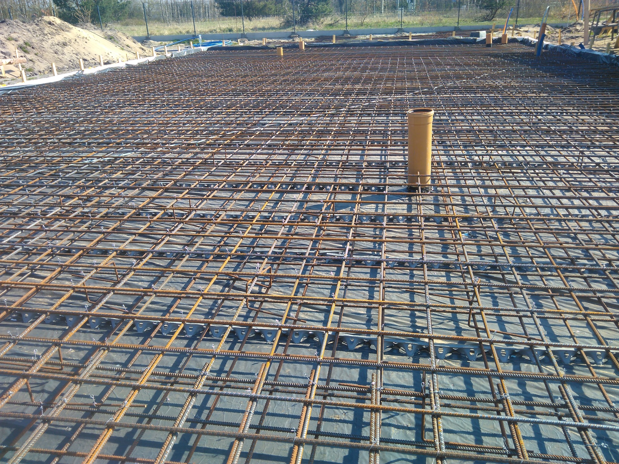 Siatka zbrojeniowa na folii, z widocznymi elementami dystansowymi i wystającymi rurami kanalizacyjnymi, przygotowana pod wylewkę fundamentu budynku.