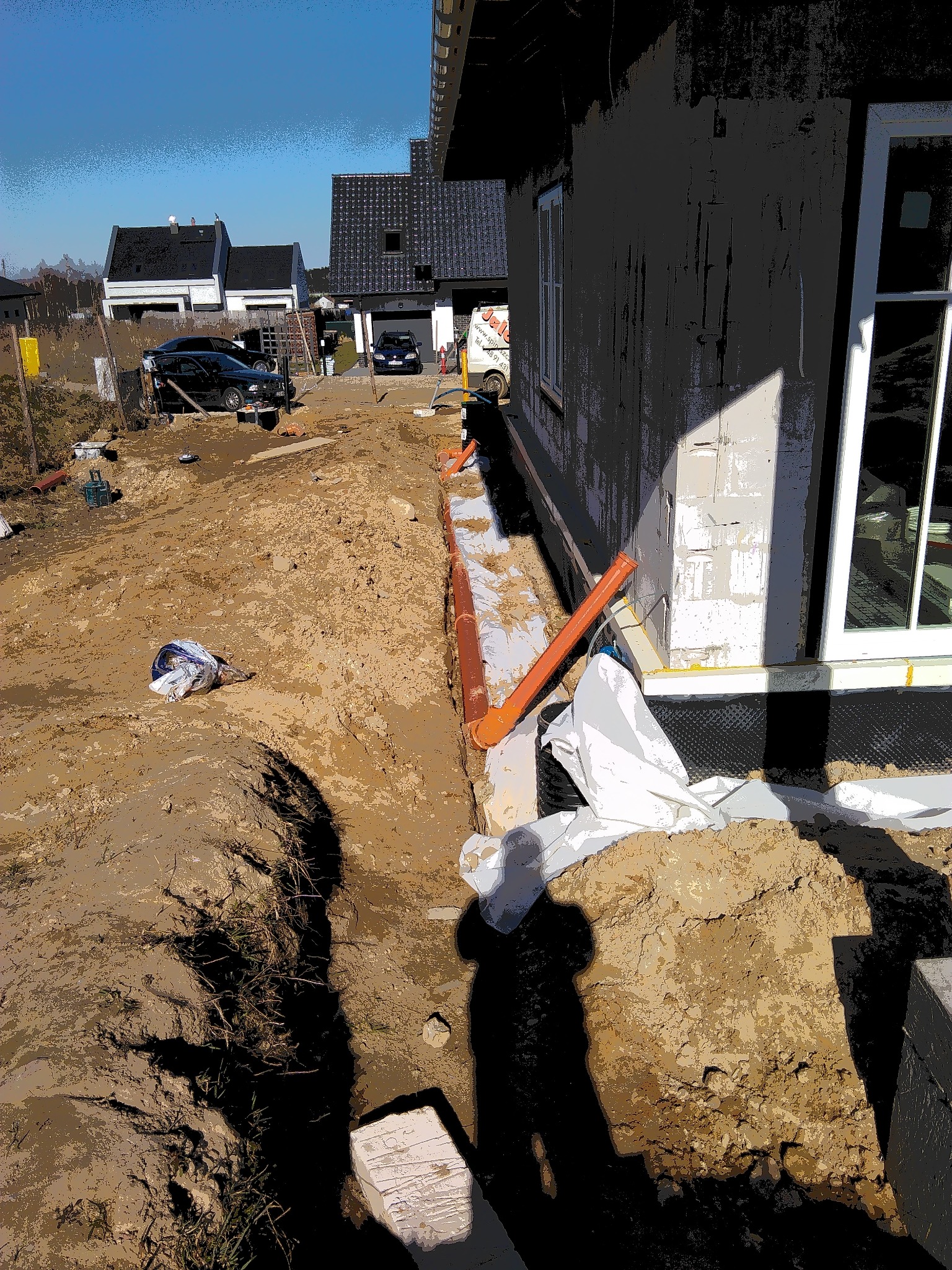 Wykop wzdłuż fundamentów domu w trakcie budowy, widoczne rury instalacyjne i folia hydroizolacyjna, w tle zabudowa jednorodzinna.