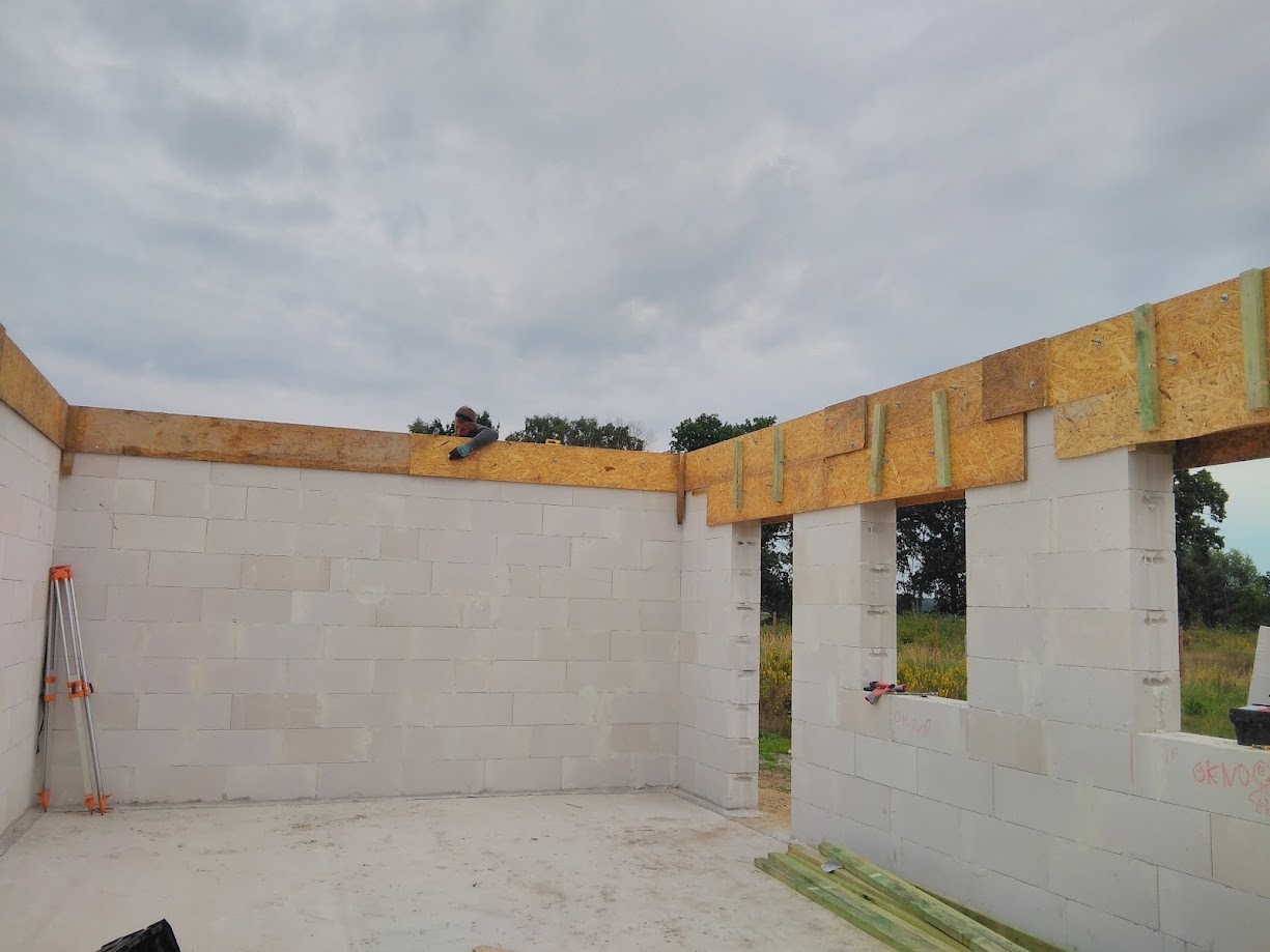 Wznoszenie ścian parterowego budynku z bloczków, widoczny pracownik montujący drewniane nadproże nad otworem okiennym, na ścianie zaznaczone okablowanie, a w tle krajobraz polny.