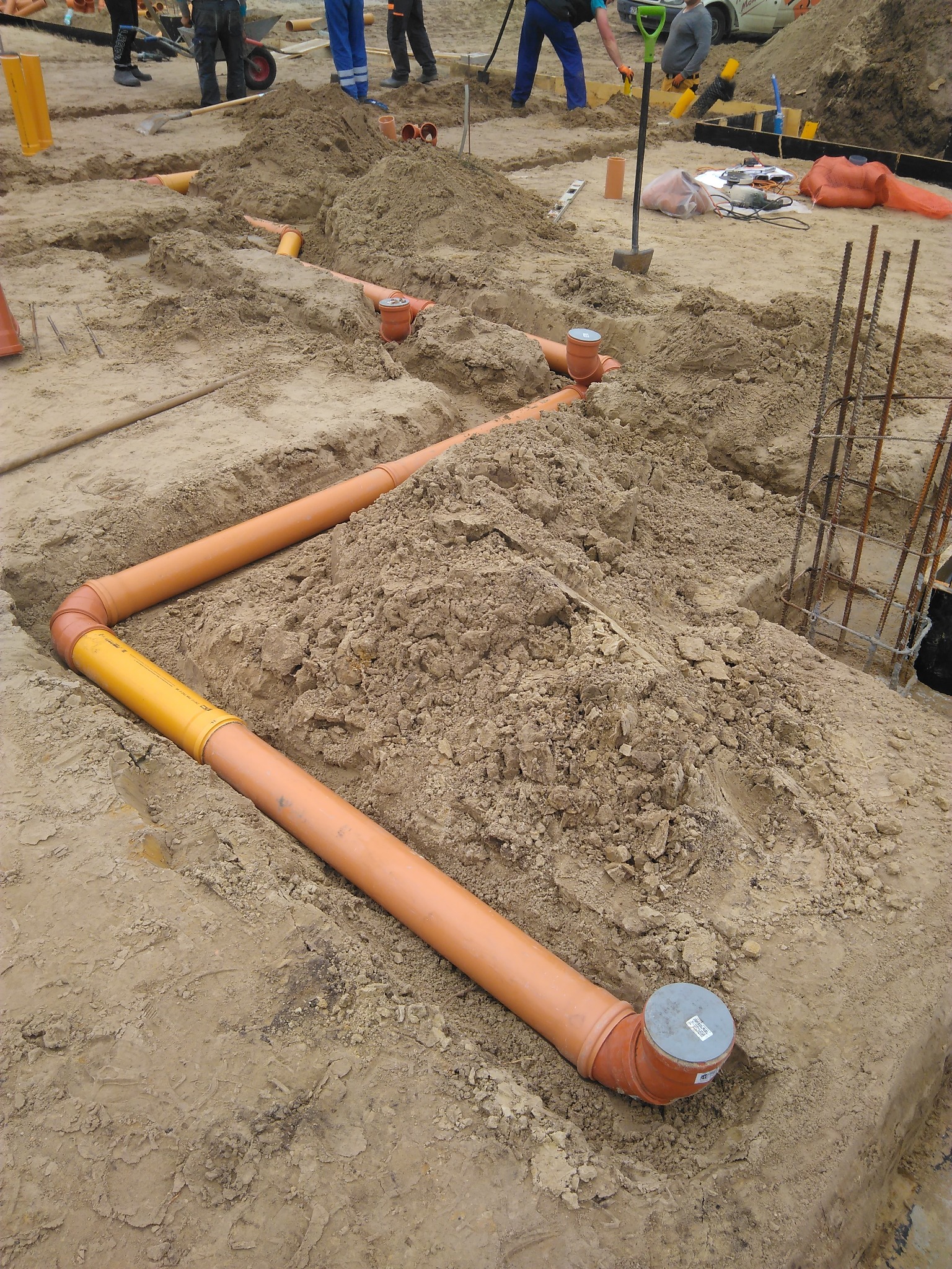 Pomarańczowe rury kanalizacyjne ułożone w wykopie ziemnym, widoczne złącza i fragmenty żółtej rury, w tle pracownicy budowlani i narzędzia.