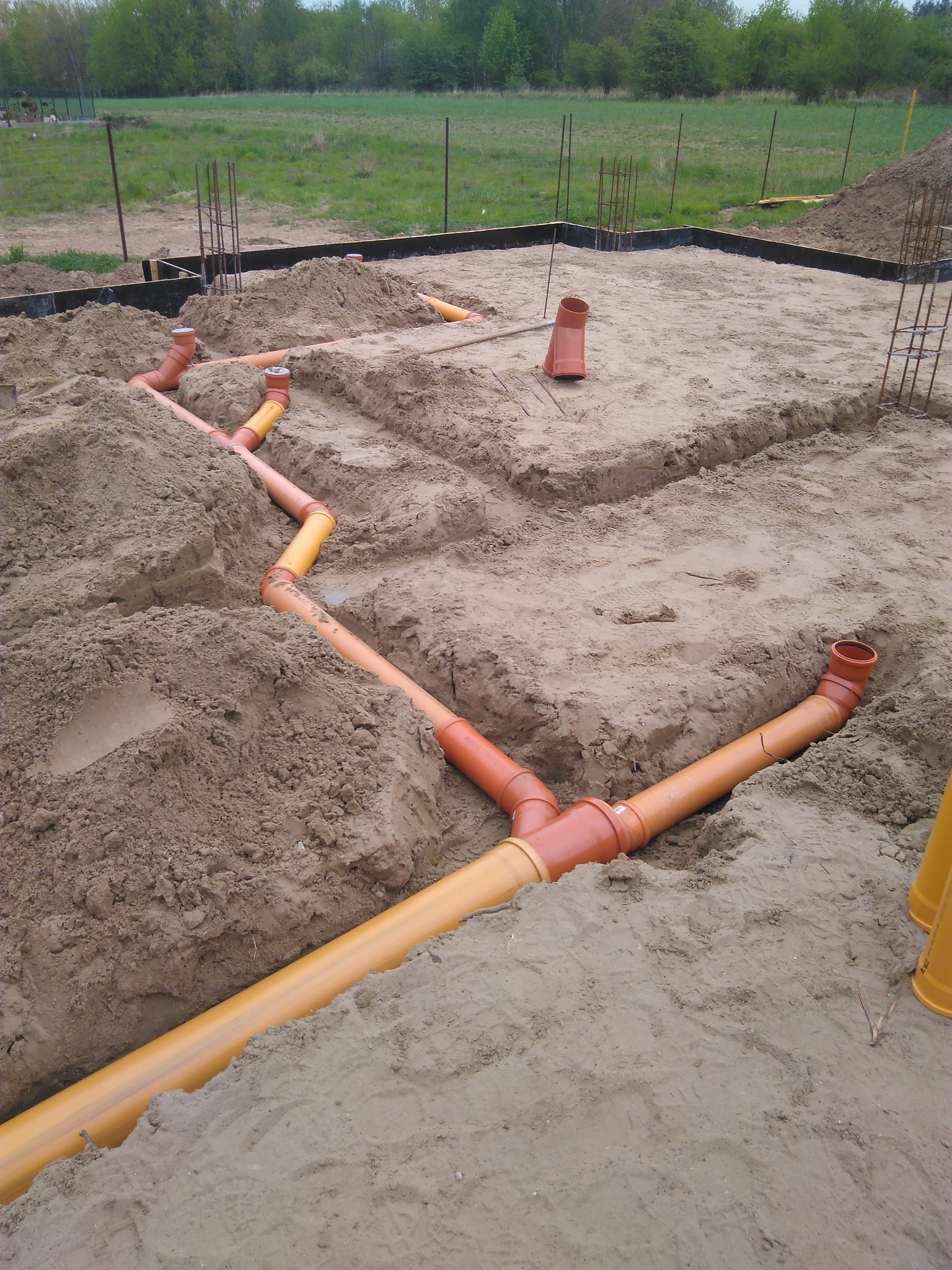 Pomarańczowe rury kanalizacyjne ułożone w wykopie fundamentowym pod budowę domu jednorodzinnego, widoczne szalunki i zbrojenie.