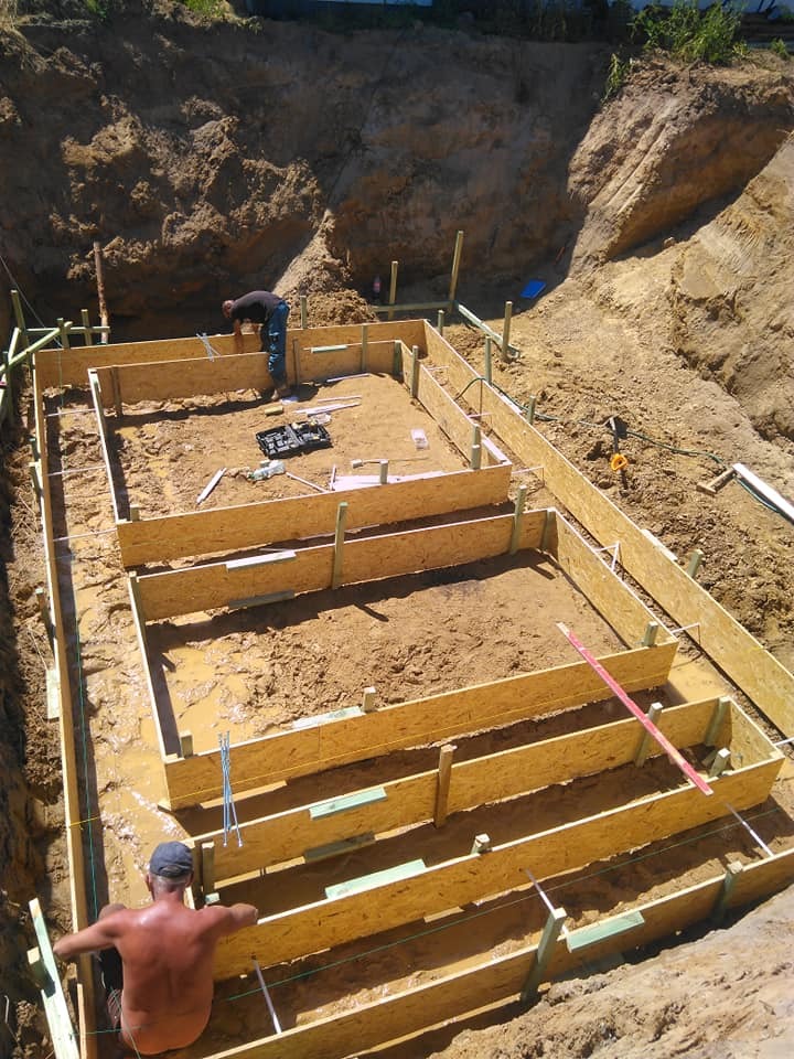 Przygotowanie wykopu pod fundamenty z deskowaniem z desek i ziemnym nasypem w tle, dwóch pracowników w trakcie prac budowlanych, narzędzia widoczne w środku wykopu.