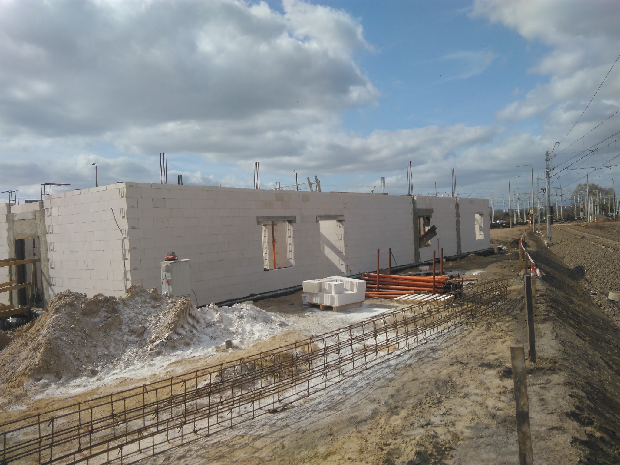 Budowa domu z białych bloczków, widoczne otwory okienne i drzwiowe, zbrojenie fundamentów w ziemi, w tle linia kolejowa i pochmurne niebo.