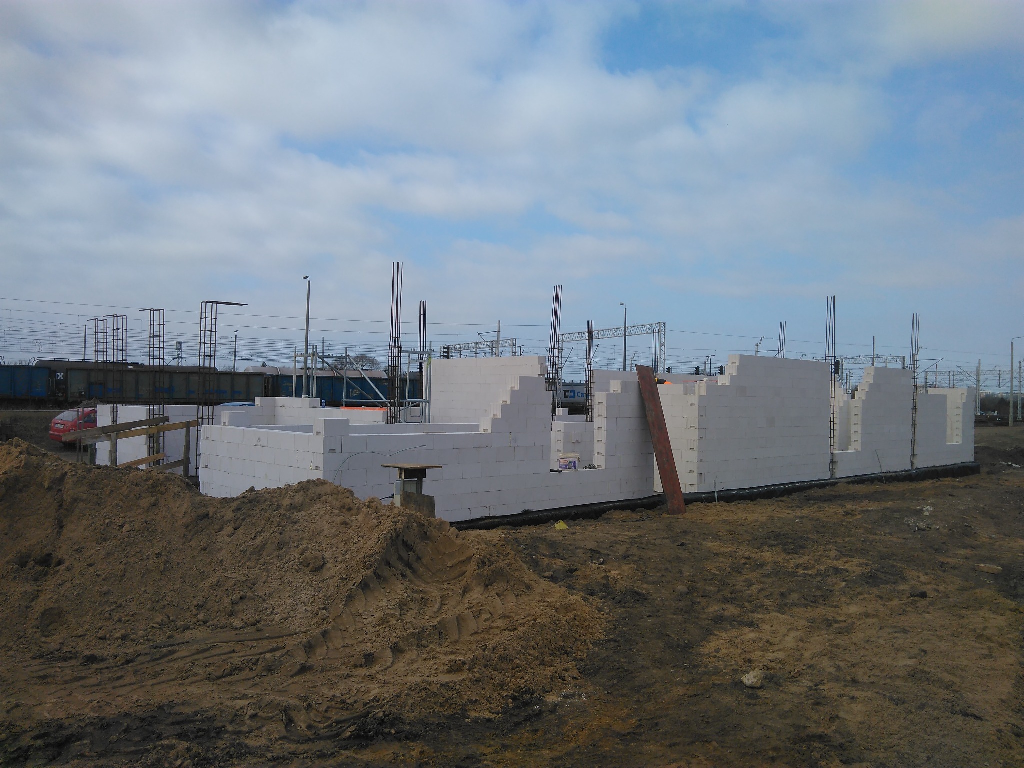 Wznoszenie ścian domu z betonu komórkowego, widoczne zbrojenie pionowe, w tle tory kolejowe i pochmurne niebo.