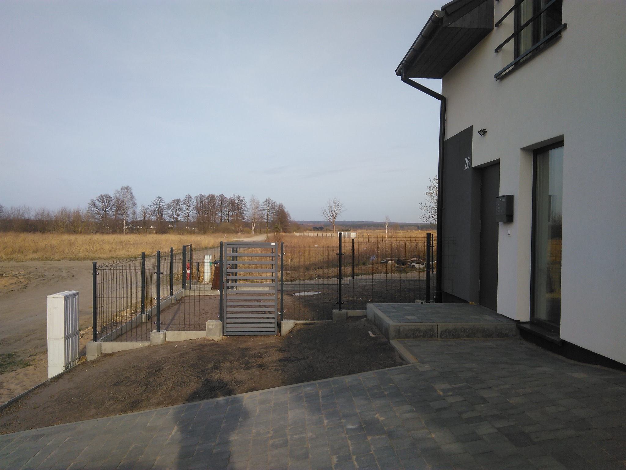 Nowoczesny dom z szarą elewacją, grafitową stolarką okienną i furtką wejściową z metalowych profili na tle jesiennego krajobrazu.