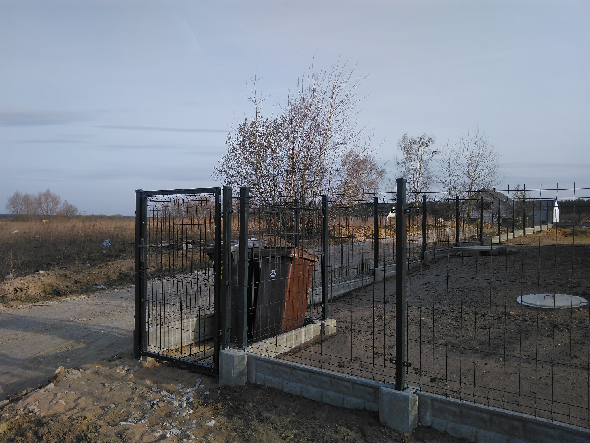 Ogrodzenie panelowe z furtką, betonowa podmurówka, brązowy kosz na śmieci wewnątrz ogrodzenia, teren w trakcie zagospodarowywania, widok na okoliczne pola i zabudowania w tle.