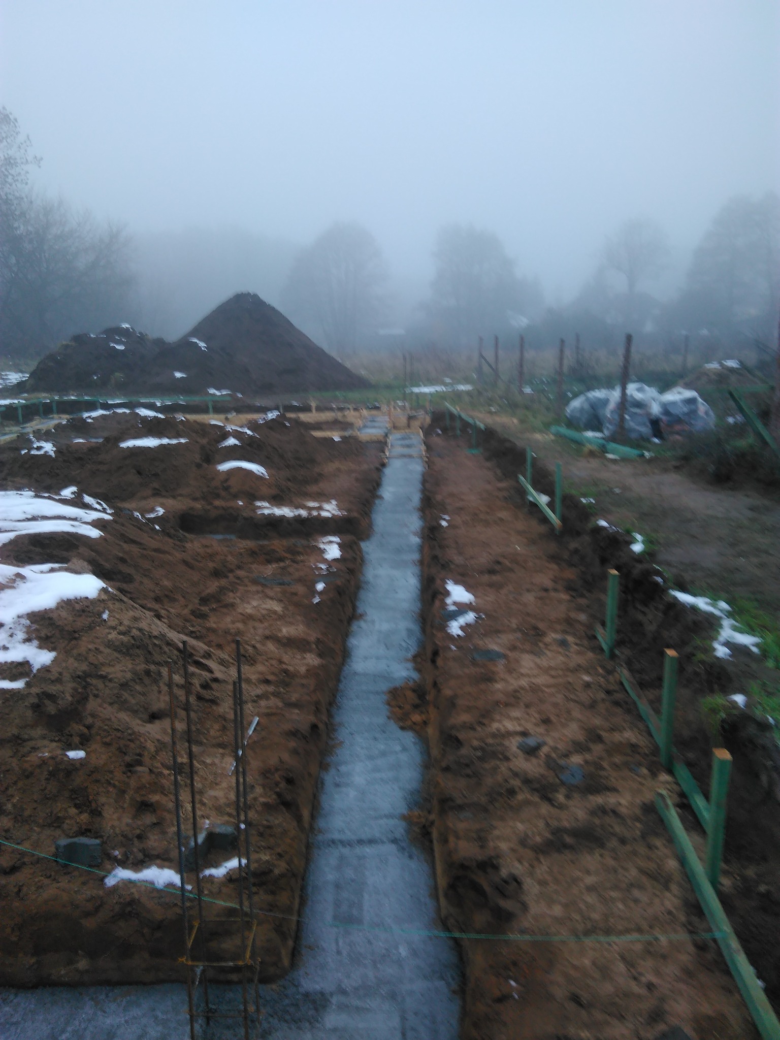 Wykop pod fundamenty, z widoczną hydroizolacją i zbrojeniem, w tle hałda ziemi i mglista sceneria.