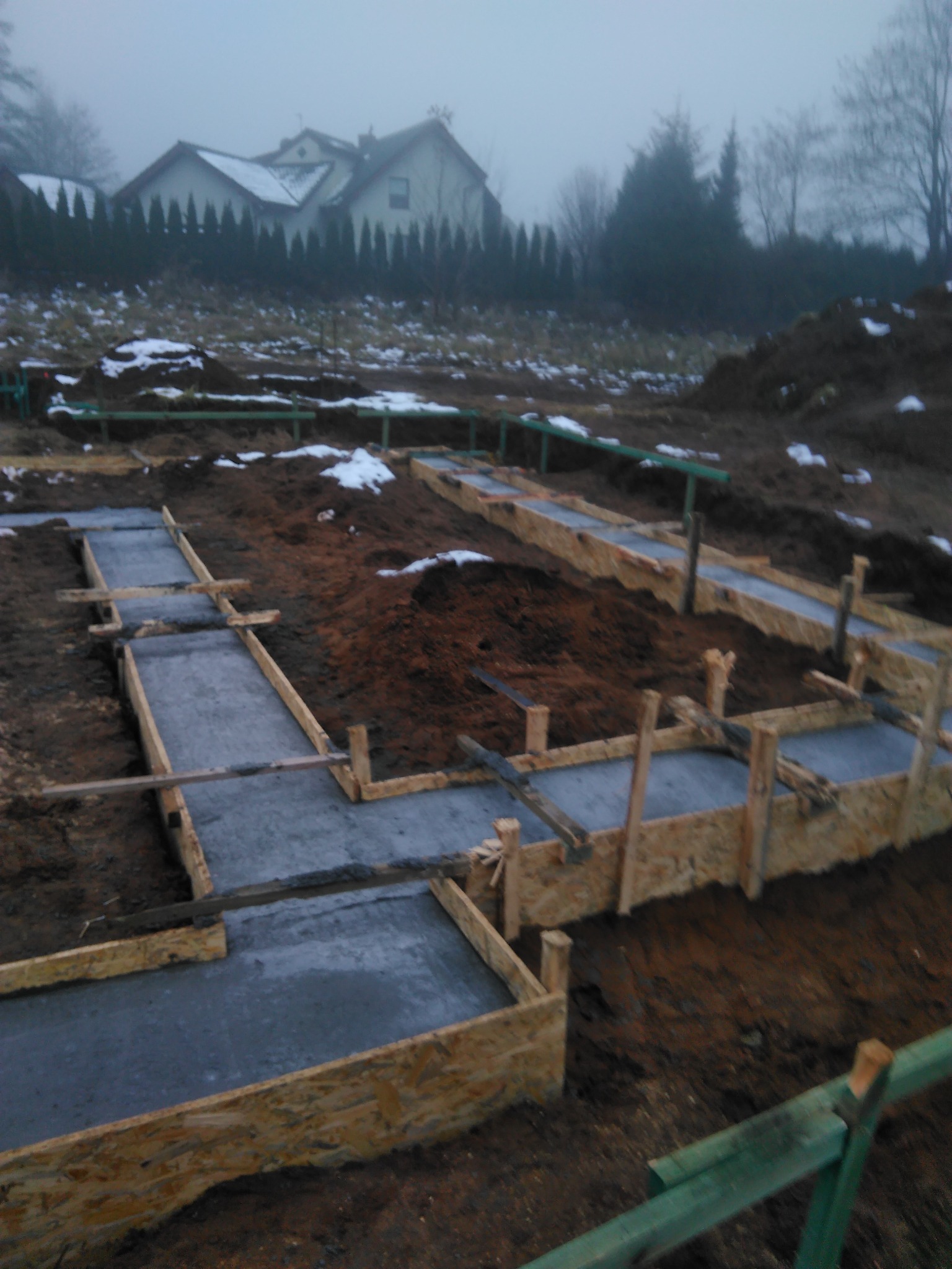 Wylane fundamenty pod budynek w drewnianym szalunku, widoczne zbrojenie i świeży beton, w tle zarys domu w zimowej scenerii.