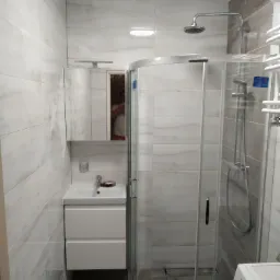 Nowoczesna łazienka z kabiną prysznicową z zaokrąglonym szkłem, umywalką z szafką w kolorze białym i dużymi, jasnymi płytkami imitującymi marmur na ścianach, dodatkowo widoczna pralka.