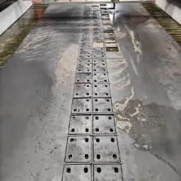 Widok z góry na stół roboczy maszyny do cięcia wodą, pokryty wodą i resztkami materiału, z widocznymi kwadratowymi płytami mocującymi z otworami.