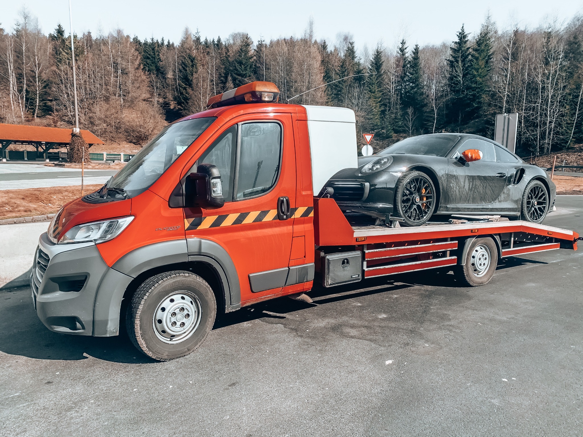 Pomarańczowa laweta z czarnym Porsche na platformie, zaparkowana na asfalcie, w tle drzewa.