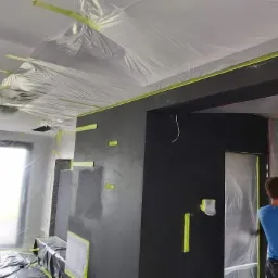 Przygotowanie pokoju do prac wykończeniowych: ściany pomalowane na ciemny kolor, folia ochronna na suficie i elementach wyposażenia, taśma malarska zabezpieczająca krawędzie, widoczna sylwetka osoby.