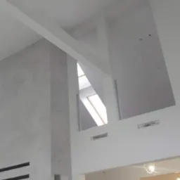 Wysokie pomieszczenie z szarą ścianą z poziomymi wnękami, białą konstrukcją z płyt kartonowo-gipsowych tworzącą wnęki i otwór okienny w dachu.