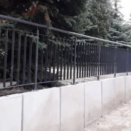 Długa, czarna, metalowa balustrada na betonowym murku oporowym, otoczona iglakami.