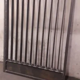 Surowa, spawana konstrukcja metalowa z pionowymi prętami i poziomą podstawą, prawdopodobnie fragment ogrodzenia lub bramy, oparta o ścianę w pomieszczeniu.