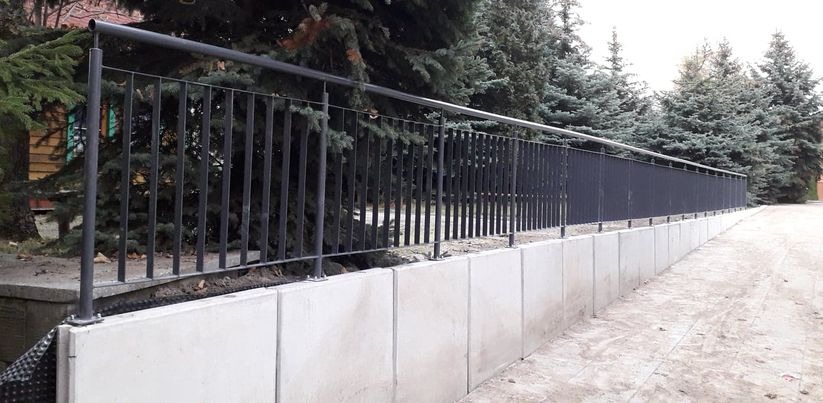 Długa, czarna, metalowa balustrada na betonowym murku oporowym, otoczona iglakami.