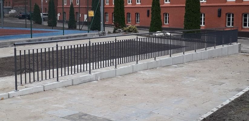 Metalowa balustrada w kolorze grafitowym na betonowej podmurówce, z widocznym placem budowy w tle i budynkiem z czerwonej cegły.