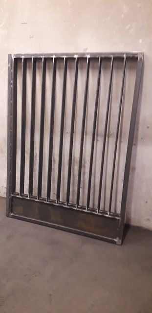 Surowa, spawana konstrukcja metalowa z pionowymi prętami i poziomą podstawą, prawdopodobnie fragment ogrodzenia lub bramy, oparta o ścianę w pomieszczeniu.