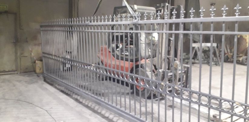 Szary metalowy płot panelowy z ozdobnymi zakończeniami w kształcie lilii, w tle wózek widłowy w hali.