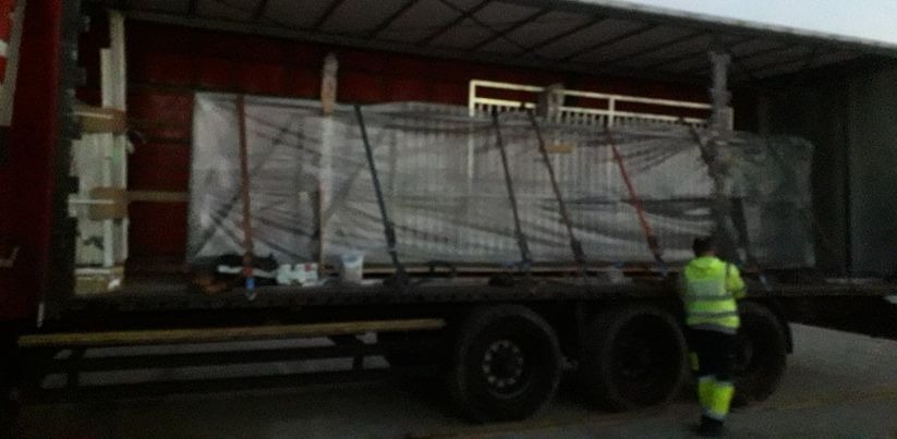 Załadunek konstrukcji stalowej na naczepę ciężarówki, zabezpieczonej pasami transportowymi i folią stretch, w tle widoczny pracownik w odblaskowej kamizelce.