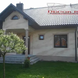 Jasna elewacja domu jednorodzinnego z dekoracyjnym wzorem w odcieniach beżu, widoczny fragment logo firmy Decor Bud na dachu, w tle zieleń ogrodu.