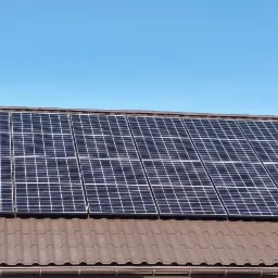 Panele słoneczne zamontowane na brązowym dachu z dachówki ceramicznej, widoczne mocowania paneli, na tle błękitne niebo.