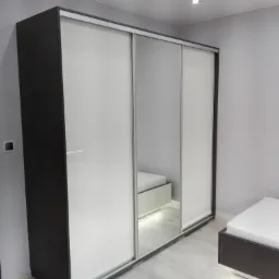 Nowoczesna szafa przesuwna z białymi frontami i ciemnobrązowymi bokami, z lustrem na środkowych drzwiach, odbijającym fragment łóżka, na tle szarej ściany i jasnej podłogi.