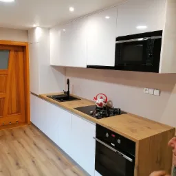 Nowoczesna kuchnia z białymi, lakierowanymi szafkami, drewnianym blatem, czarnym zlewem i płytą gazową z czajnikiem w kwiaty, oświetlona punktowymi lampami sufitowymi.