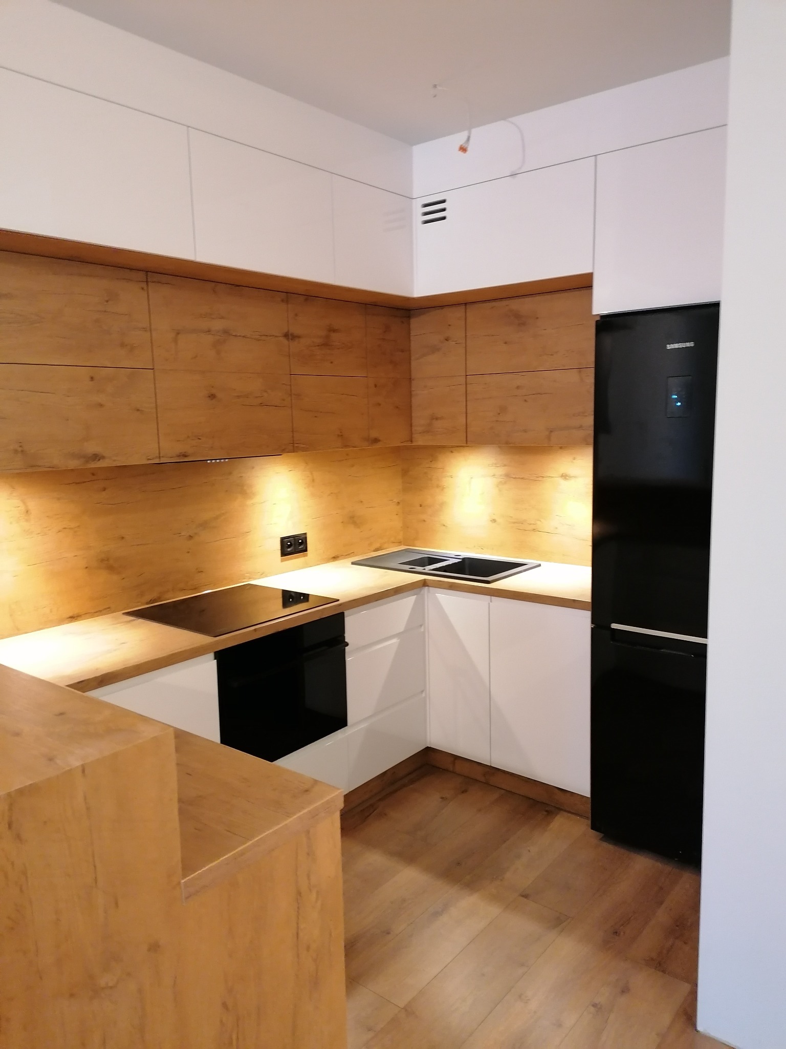 Nowoczesna kuchnia z białymi szafkami i drewnianymi blatami, czarna lodówka Samsung, płyta indukcyjna i oświetlenie podszafkowe.
