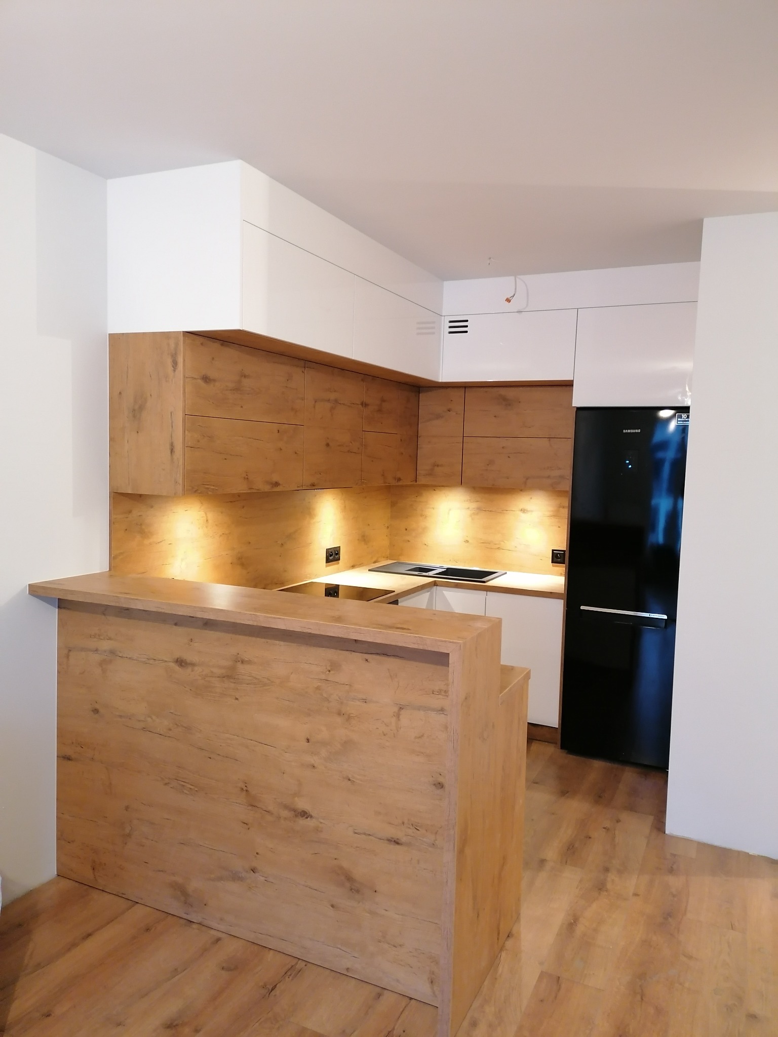 Nowoczesna kuchnia z drewnianymi frontami szafek górnych i barkiem, białe szafki dolne i czarna lodówka w zabudowie, oświetlenie LED pod szafkami.