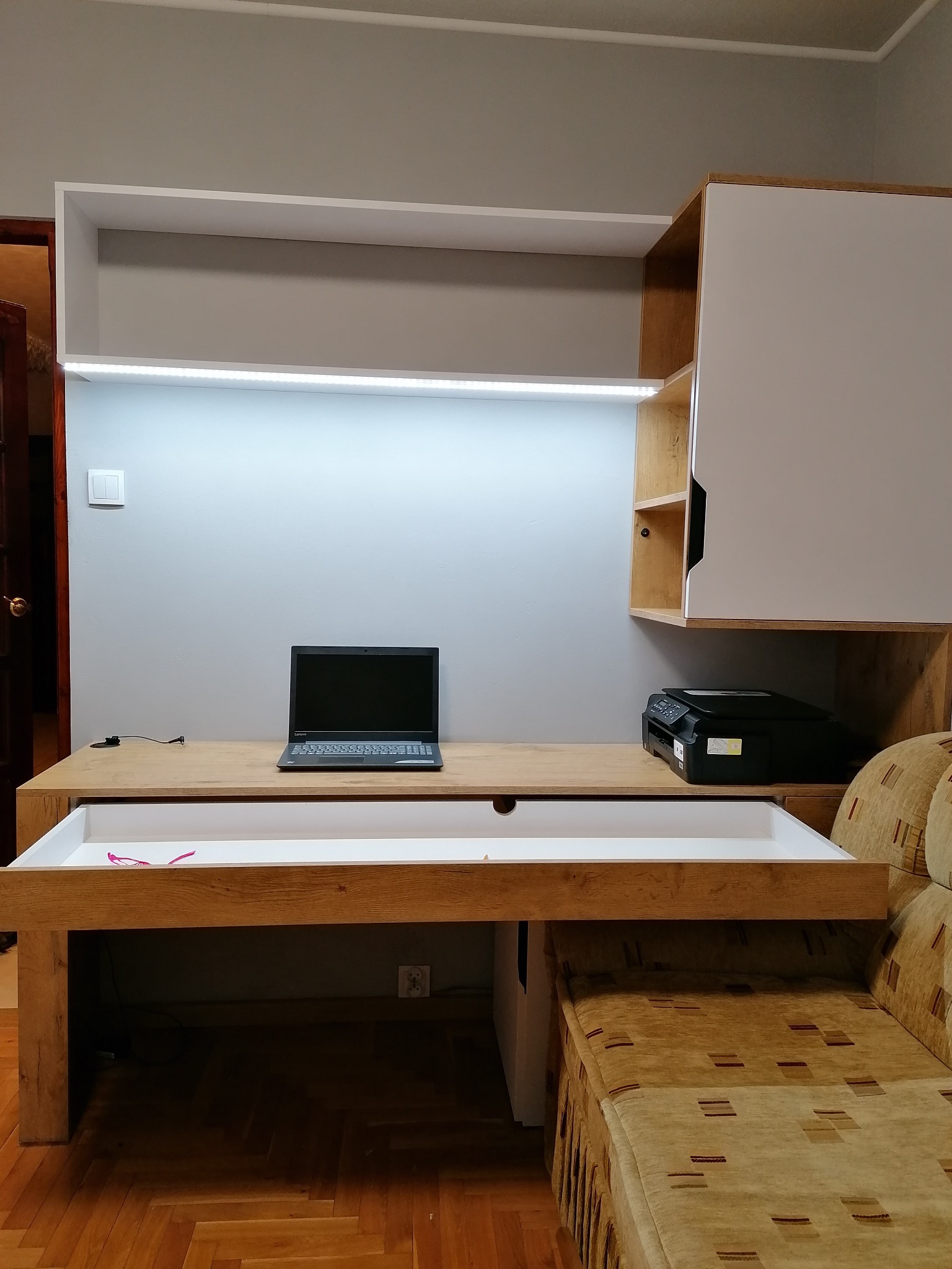 Drewniane biurko z wysuwaną szufladą, nad nim szafka z oświetleniem LED, na blacie laptop i drukarka, obok sofa w brązowych odcieniach.