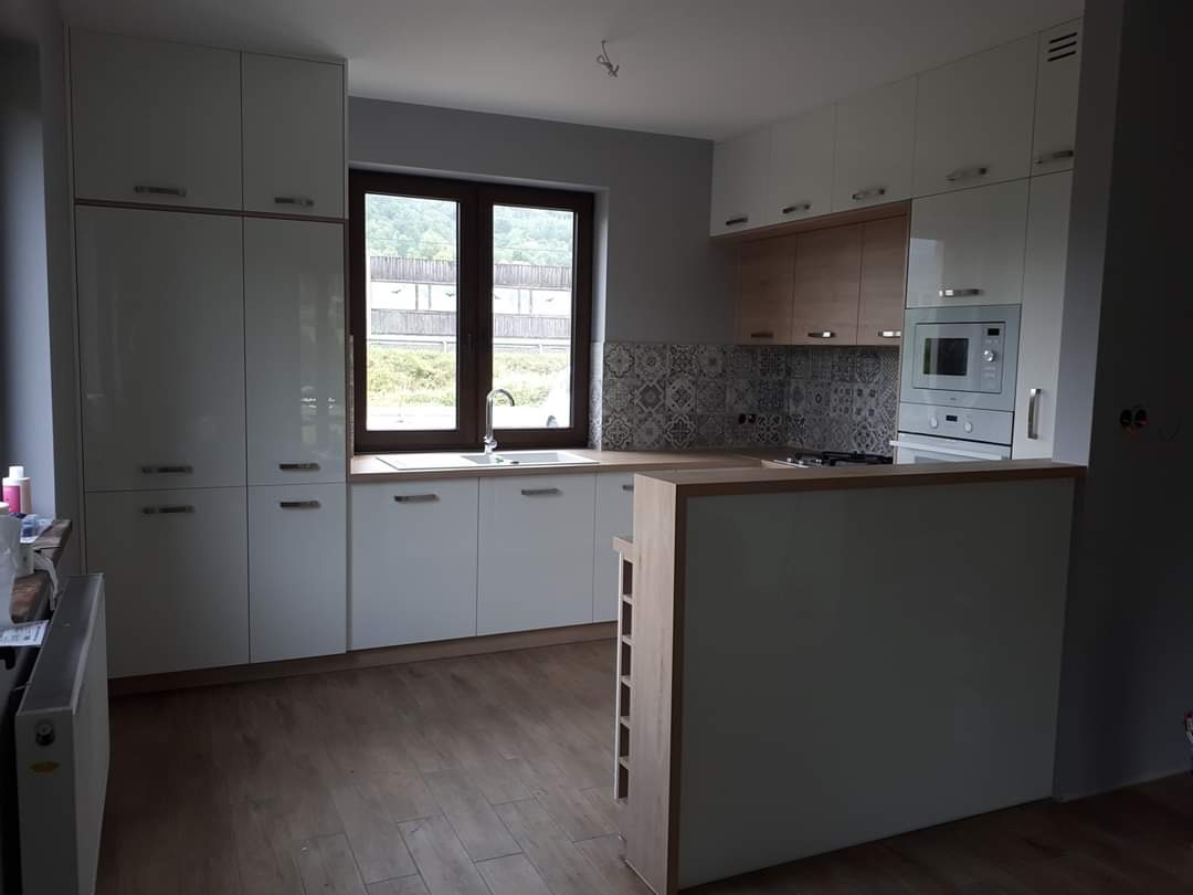 Nowoczesna kuchnia z białymi, błyszczącymi frontami szafek, drewnianym blatem i wzorzystą mozaiką nad blatem roboczym, widok na okno z widokiem na zieleń.