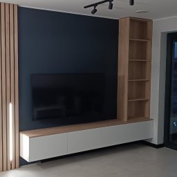 Meble Olejniczak - Nowoczesna zabudowa RTV z drewnianymi lamelami z podświetleniem LED, szafką RTV i regałem w odcieniach drewna, na tle ciemnej ściany i przesuwnych drzwi balkonowych.