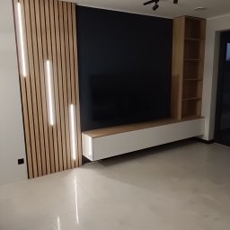Meble Olejniczak - Nowoczesny salon z podświetlanymi lamelami na ścianie, szafką RTV i regałem w odcieniach drewna. Czarna ściana za telewizorem, minimalistyczne oświetlenie sufitowe.