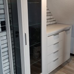 Meble Olejniczak - Biała szafa witrynowa z szufladami i przeszkleniami na tle ceglanej ściany i drewnianej podłogi. Mebel na metalowych nóżkach, wkomponowany w przestrzeń pod skosem.