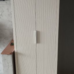 Meble Olejniczak - Biała szafa dwudrzwiowa z ryflowanymi frontami, minimalistycznymi uchwytami i smukłymi nóżkami, ustawiona na podłodze z paneli w odcieniach szarości.