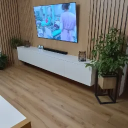 Biała szafka RTV pod telewizorem na tle ściany z drewnianych paneli. Na szafce dekoracje i zegar elektroniczny. Parkiet z paneli drewnopodobnych.