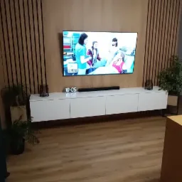 Nowoczesny salon z drewnianą ścianą z lamelami. Biała szafka RTV pod telewizorem, rośliny doniczkowe po bokach. Drewniana podłoga.