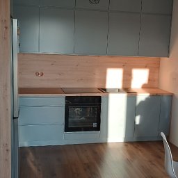 Meble Olejniczak - Kompletna kuchnia z szarymi szafkami i drewnianym blatem, oświetlona naturalnym światłem słonecznym, z widoczną lampą wiszącą w stylu industrialnym.