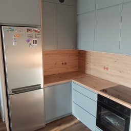 Meble Olejniczak - Narożna, szara kuchnia na wymiar z drewnianym blatem i srebrną lodówką. Nad blatem drewniana ściana. Wisząca lampa w stylu industrialnym. Całość w nowoczesnym stylu.