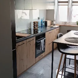 Nowoczesna kuchnia z szarymi szafkami górnymi i drewnopodobnymi dolnymi. Czarny blat, indukcja, piekarnik. Nad blatem białe płytki. Na podłodze szare płytki imitujące kamień.