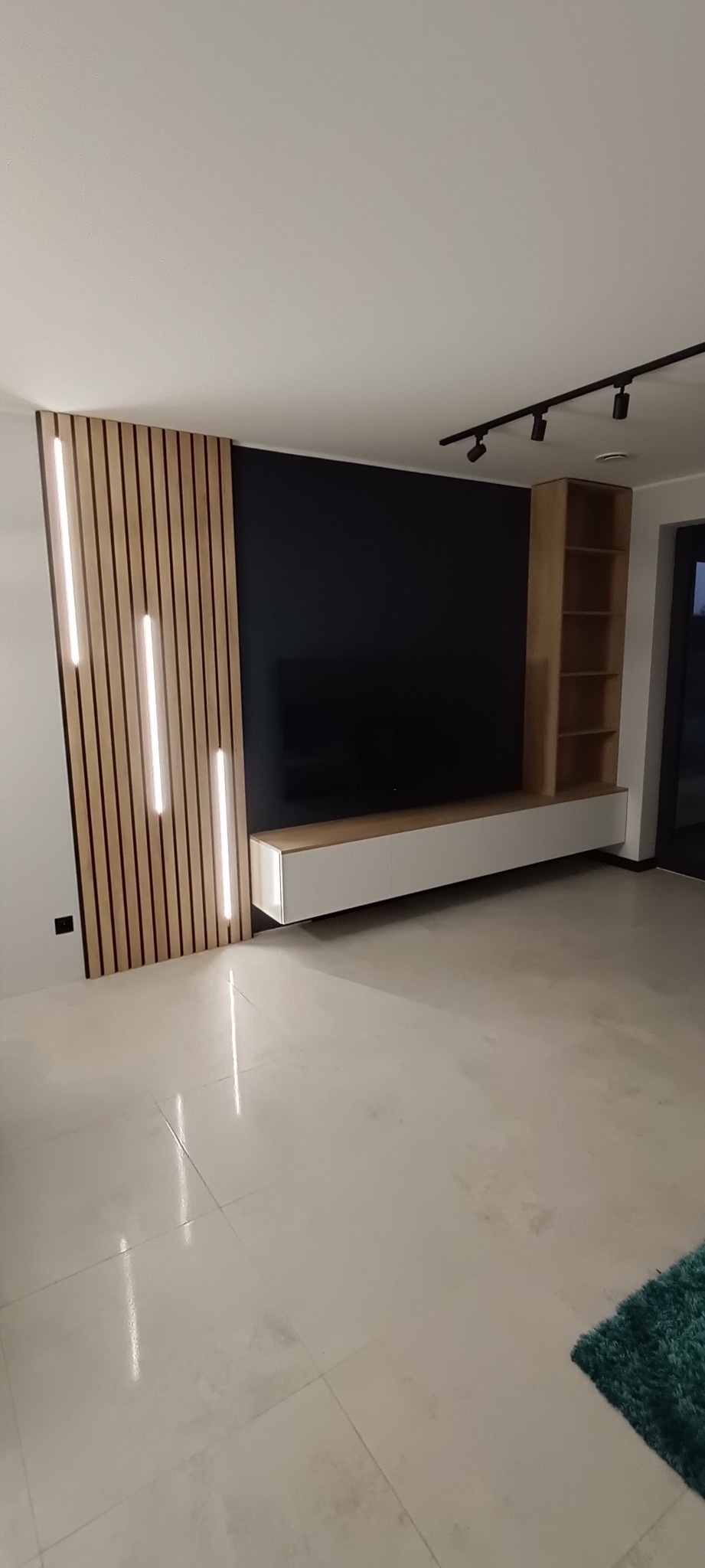 Nowoczesny salon z podświetlanymi lamelami na ścianie, szafką RTV i regałem w odcieniach drewna. Czarna ściana za telewizorem, minimalistyczne oświetlenie sufitowe.