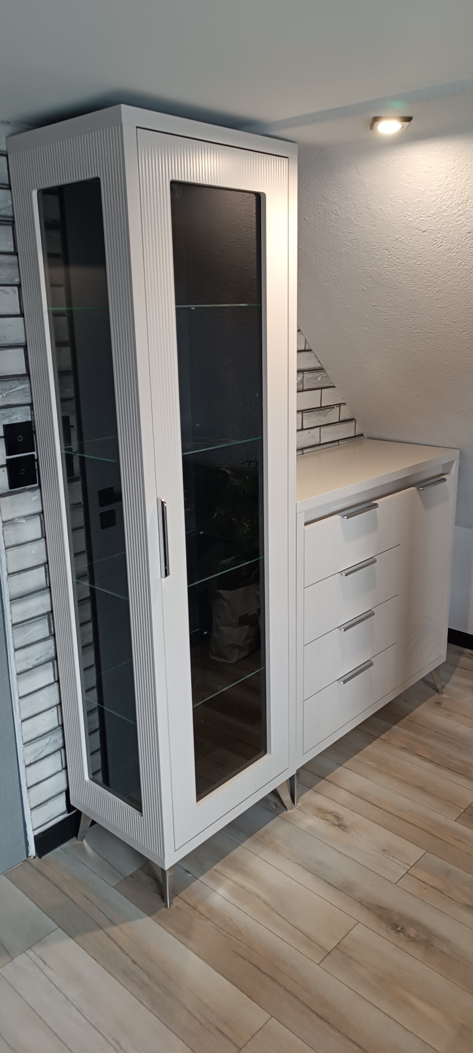 Biała szafa witrynowa z szufladami i przeszkleniami na tle ceglanej ściany i drewnianej podłogi. Mebel na metalowych nóżkach, wkomponowany w przestrzeń pod skosem.