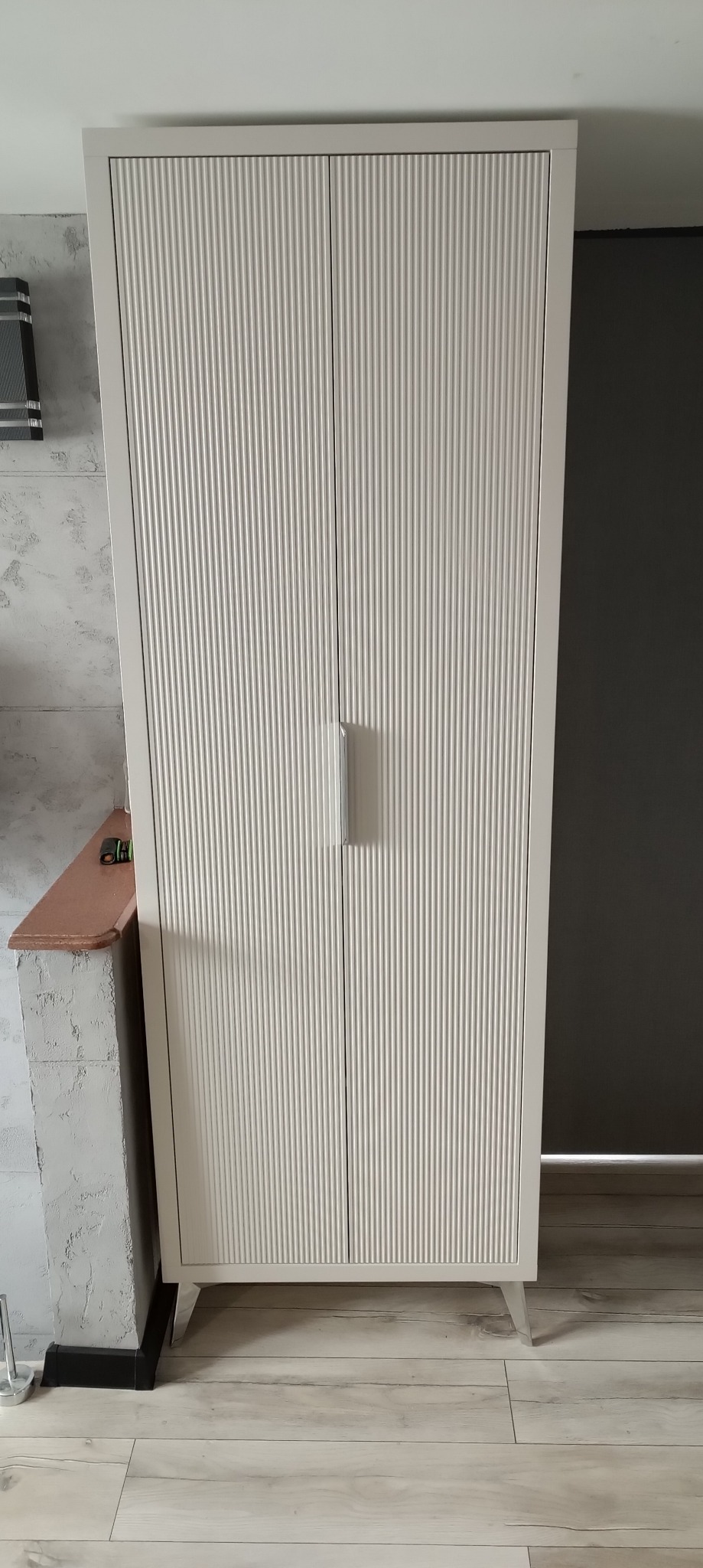 Biała szafa dwudrzwiowa z ryflowanymi frontami, minimalistycznymi uchwytami i smukłymi nóżkami, ustawiona na podłodze z paneli w odcieniach szarości.