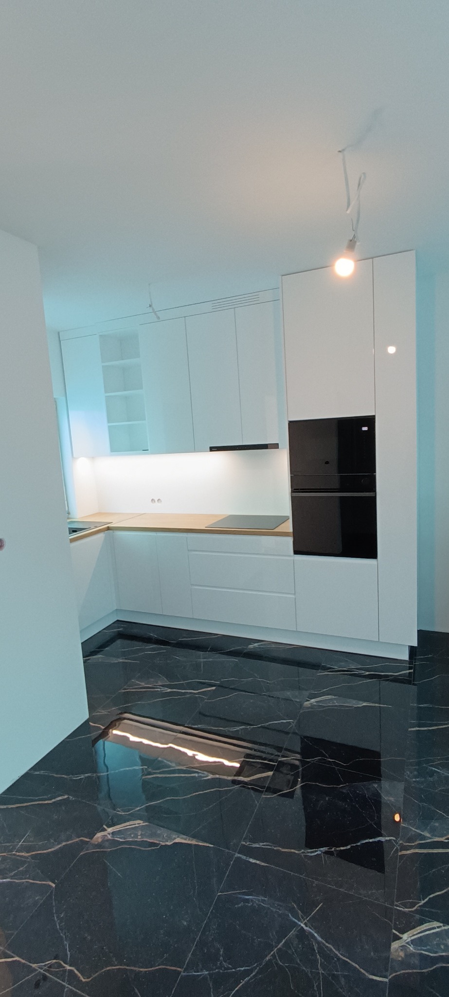 Nowoczesna, biała kuchnia z drewnianym blatem i czarnym piekarnikiem, odbijająca się w błyszczącej, czarnej podłodze. Minimalistyczny design, jasne oświetlenie.