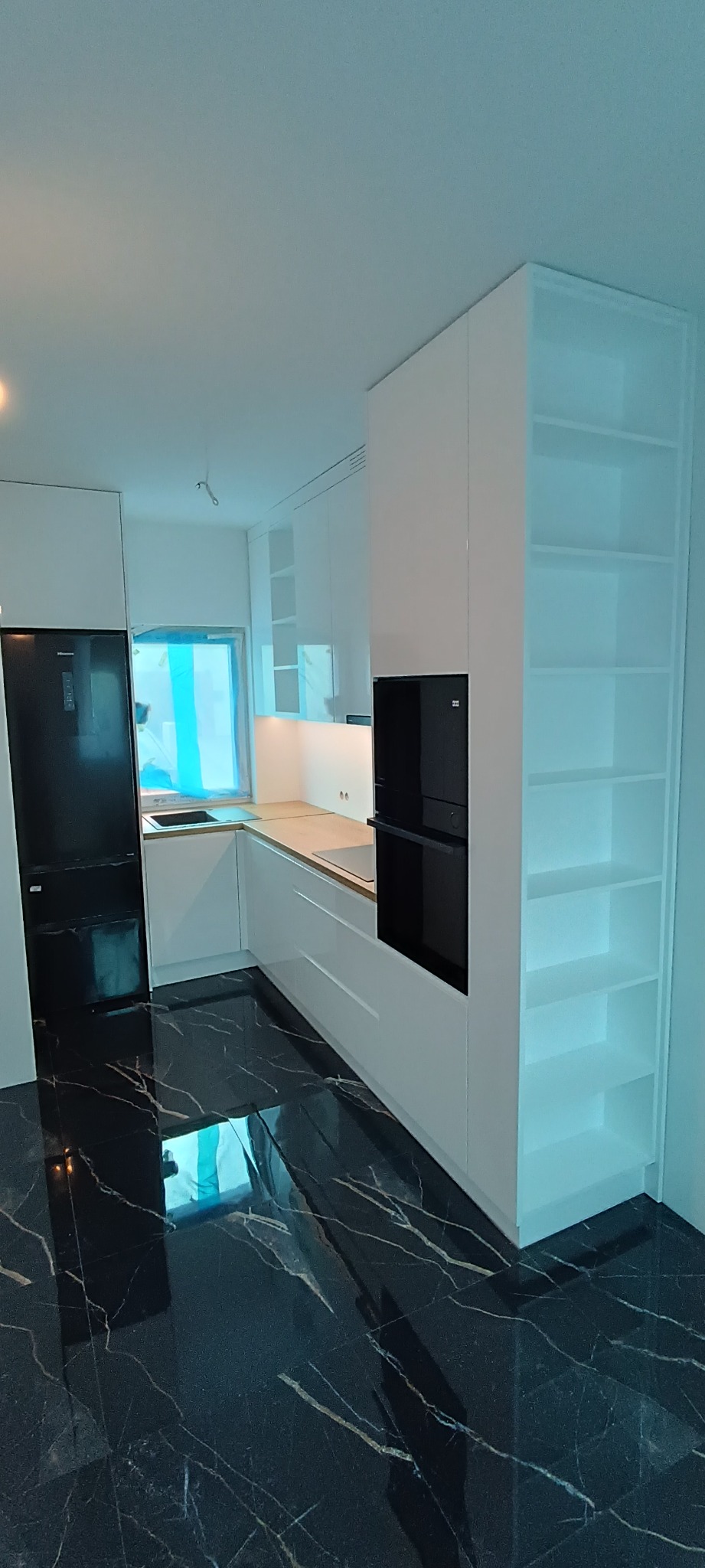 Biała kuchnia z czarnym blatem i sprzętem AGD. Widoczne szafki, lodówka, piekarnik w zabudowie i otwarty regał. Minimalistyczny design i połyskująca podłoga.